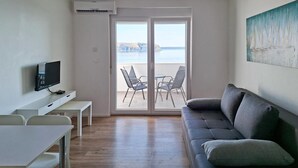 A modern living room with a flat-screen TV, a sofa, a dining table, and chairs. There is a sliding glass door leading to an outdoor area with a view of the sea and cliffs. - Lovely apartment in Rtina with WiFi (Rtina)