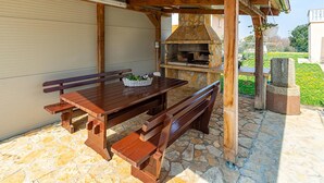 An outdoor patio with a stone-tiled floor, a wooden table with benches, a stone fireplace, and a hanging light fixture.