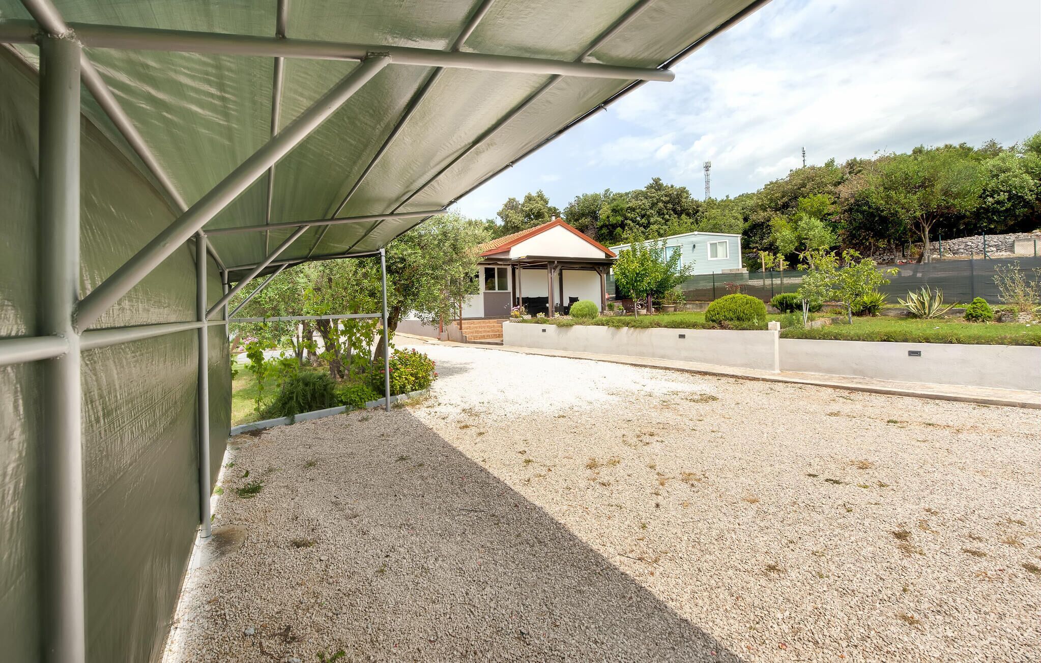 A covered outdoor area with a gravel ground, a metal fence, and a small house in the background.