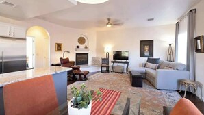 Una sala de estar moderna con un sofá, una mesa de centro y un televisor. Hay una zona de cocina con un refrigerador y alacenas.