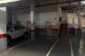 Un parking avec une voiture blanche garée dans un espace désigné, plusieurs portes de garage et un escalier menant à un niveau supérieur.