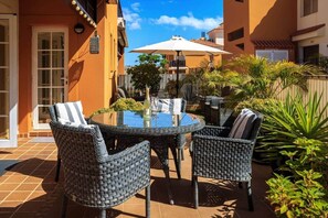 Une terrasse extérieure avec une table ronde et des chaises en osier, entourée de plantes en pot et d’un parasol blanc.
