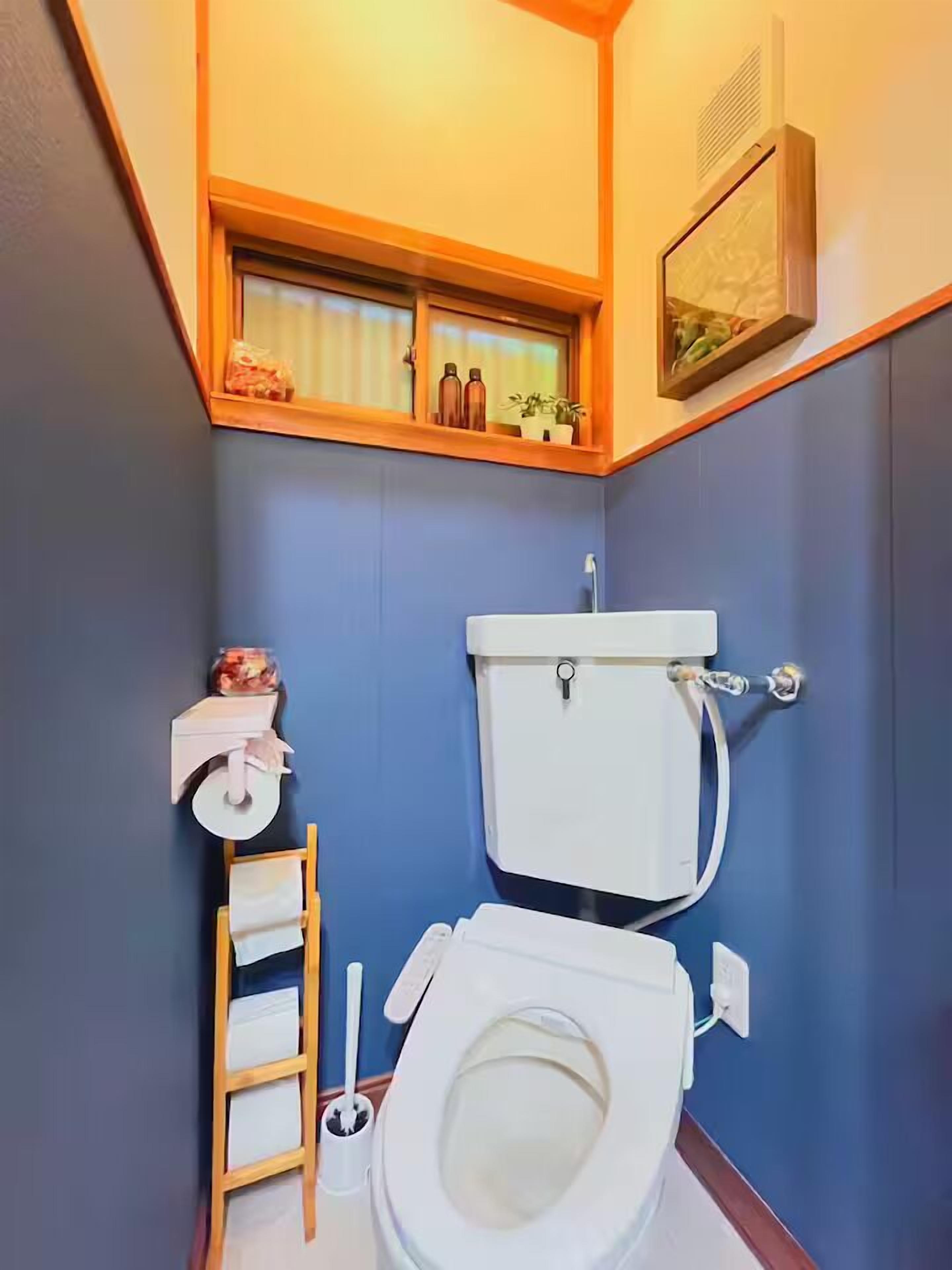 A compact bathroom with a toilet, a small shelf with toiletries, a window above the toilet, and a ladder for storage.