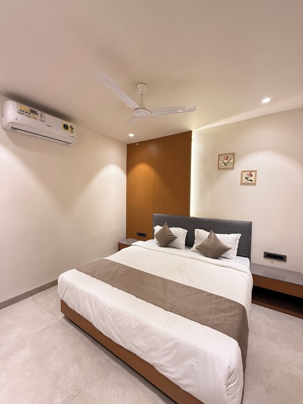 A modern bedroom with a bed, a ceiling fan, an air conditioning unit, a nightstand, and a wall-mounted shelf. - Hotel vimlayatan (Ujjain)
