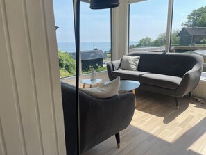 A modern living room with a grey sofa, a small round table, and a view of the sea through large windows.