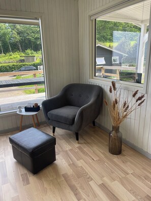 A room with a grey armchair, a small side table, a black ottoman, a vase with dried plants, and a large window with a view of greenery and a building outside.