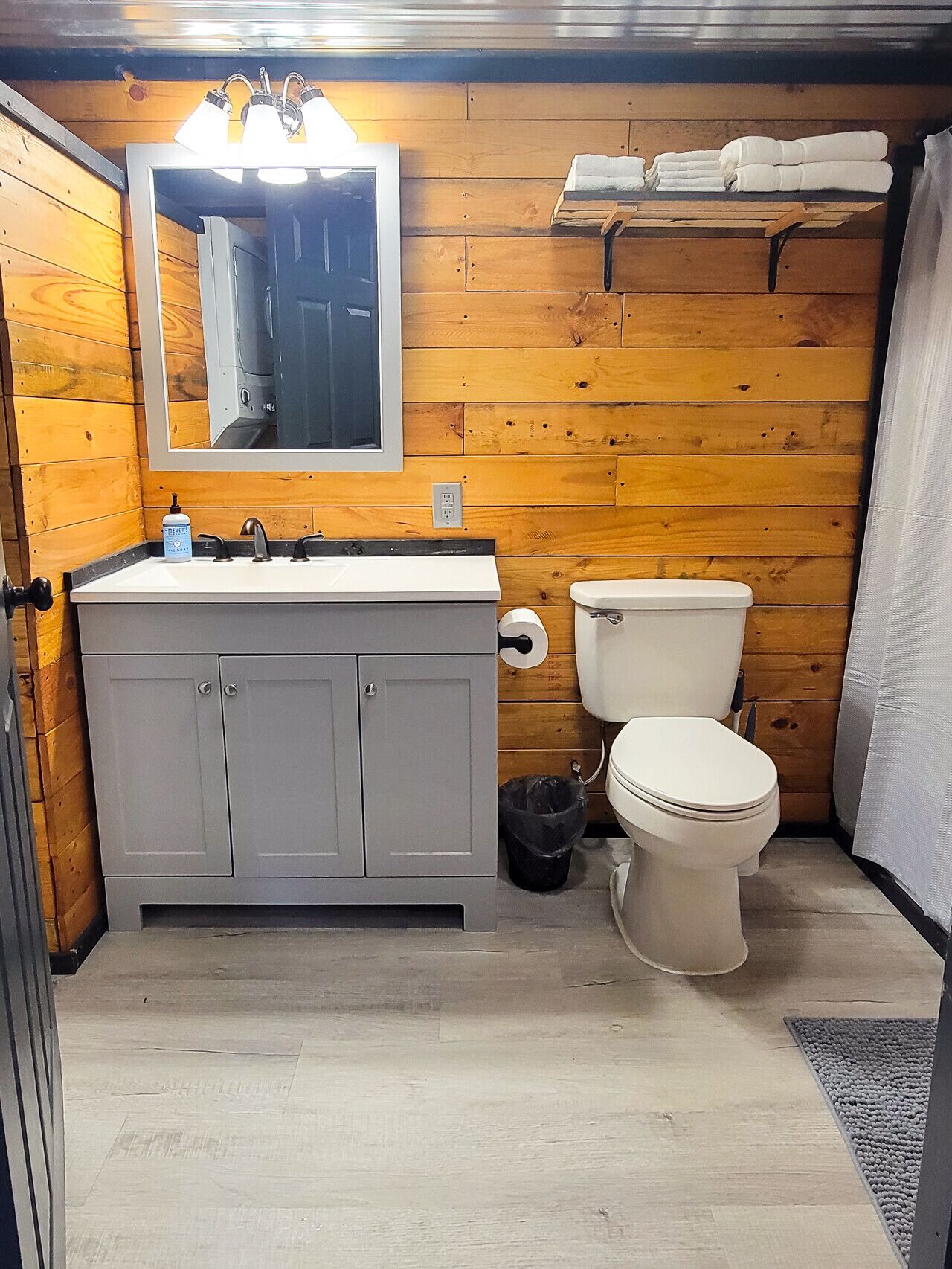 A compact bathroom with a wooden wall, a white sink with a mirror above it, a toilet, and a small shelf with towels.