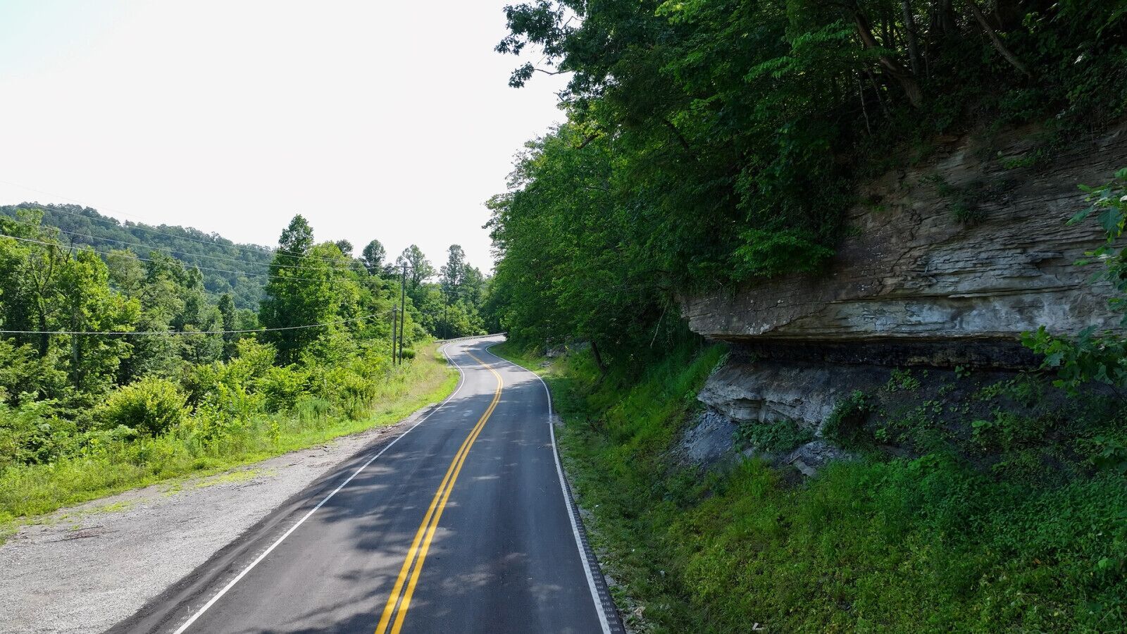 A winding road with a double yellow line, surrounded by lush greenery and a rocky cliff.