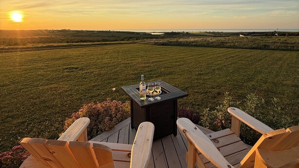 A wooden deck with two Adirondack chairs facing a grassy field. A small table with a bottle and glasses is placed in the center. - Fun for the whole Family with a beautiful view! (French River)