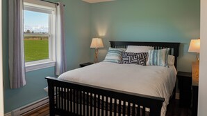 A bedroom with a wooden bed, two bedside tables with lamps, a window with curtains, and a view of a green field. - Fun for the whole Family with a beautiful view! (French River)