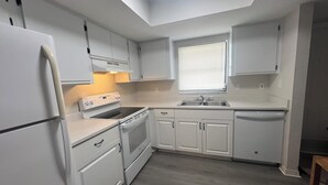 Eine kleine Küche mit weißen Schränken, einem Backofen, einem Kühlschrank und einem Fenster mit Jalousien.