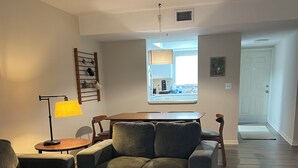Ein modernes Wohnzimmer mit einer Couch, einem Couchtisch und einer Lampe. Durch ein Fenster ist eine Küche erkennbar.