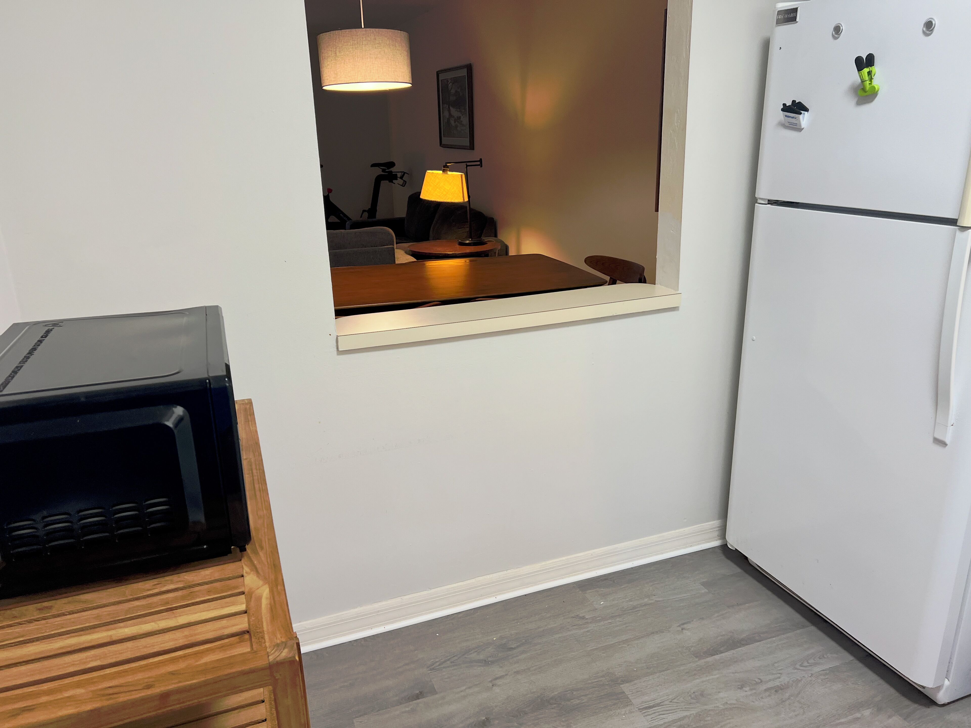 Uma cozinha compacta com micro-ondas sobre bancada de madeira, frigorífico branco e zona de refeições com mesa e cadeiras.