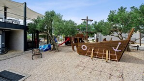 Une aire de jeux dotée d’une structure ludique en forme de bateau, d’un espace de repos couvert et d’un sol en gravier.