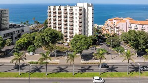 A high-rise hotel with multiple balconies, a parking lot, and a view of the ocean.