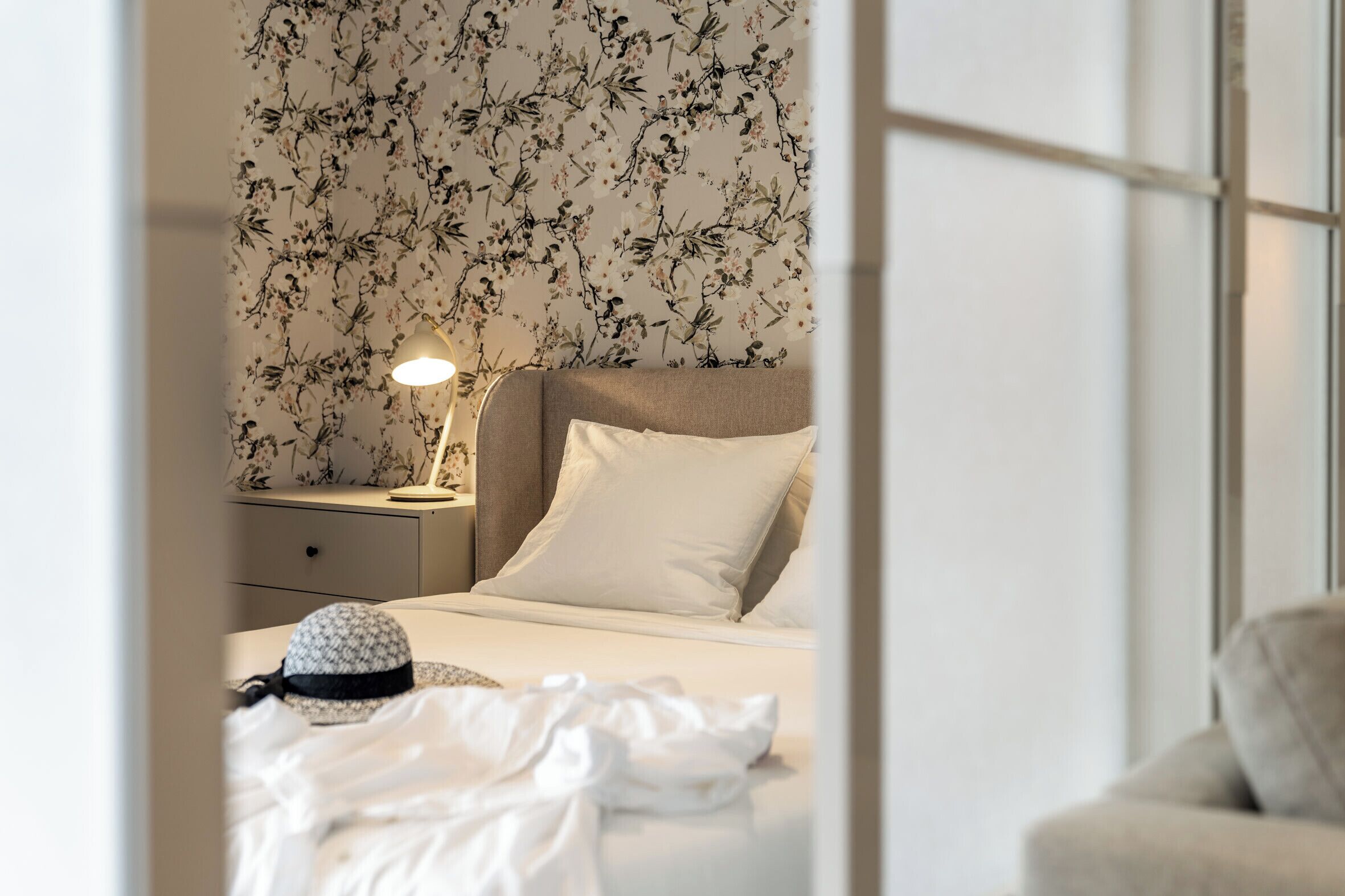 Un lit bien fait, avec une literie blanche, une lampe de chevet et un papier peint à motifs floraux.
