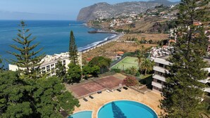 A resort with two swimming pools, a tennis court, and a view of the ocean and hills.