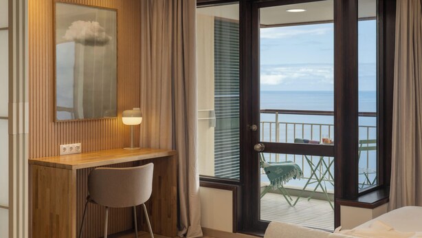 A hotel room with a wooden desk, a chair, a bed, a balcony with a view of the sea, and a large window.