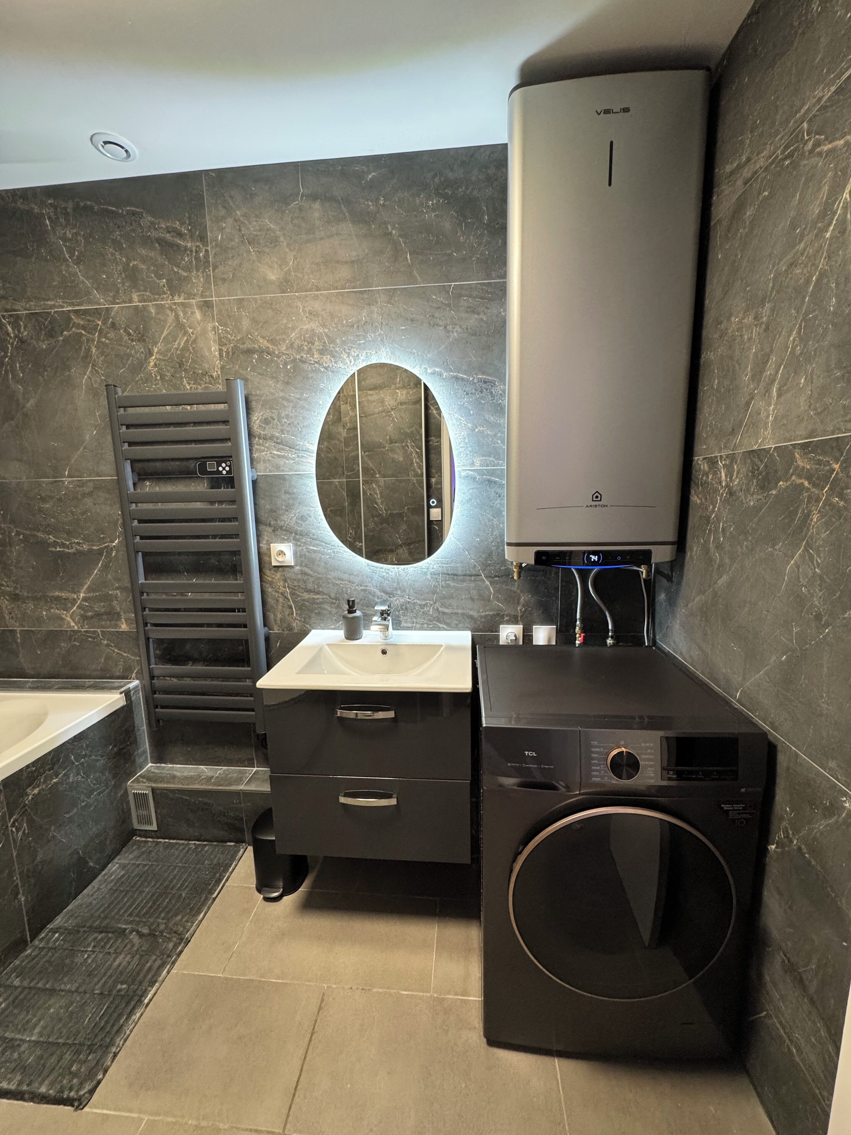 Een moderne badkamer met een ingebouwde verwarming, een wastafel, een spiegel, een handdoekhouder en een wasmachine.