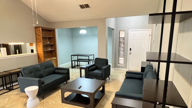 A modern living room with dark sofas, a coffee table, and a white side table. There is a wooden bookshelf and a dining table with chairs in the background.