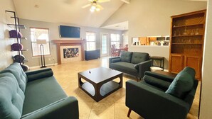 A spacious living room with a fireplace, a ceiling fan, and a kitchen area in the background.