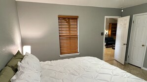 A bedroom with a large bed, a window with blinds, and a door leading to another room.