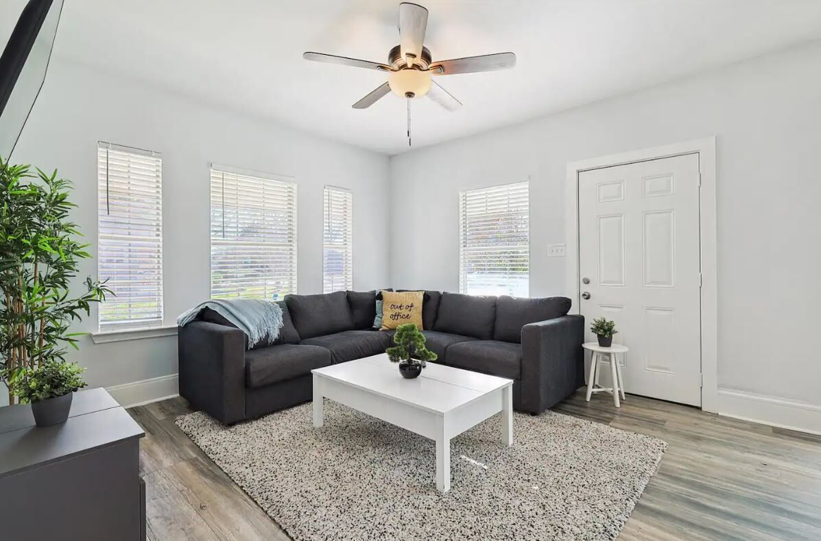 A modern living room with a dark gray sofa, a white coffee table, and a ceiling fan. There are three windows with blinds, a white door, and a small white stool with a plant.