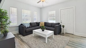 A modern living room with a dark gray sofa, a white coffee table, and a ceiling fan. There are three windows with blinds, a white door, and a small white stool with a plant.