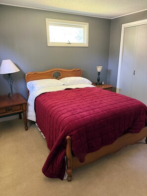 En pænt redt seng med træramme og et rødt tæppe, et sengebord med en lampe, et lille bord med et ur og et garderobeskab.
