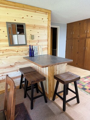 Une cuisine avec un mur en bois, un plan de travail en marbre et un meuble. On y trouve deux tabourets et une chaise.