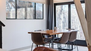 Un espace repas moderne avec une table en bois et des chaises noires, de grandes fenêtres offrant une vue sur les arbres et une poutre en bois au plafond.