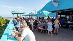 A crowded outdoor dining area with a long table and umbrellas, overlooking a body of water. - Vacation Destination (Myrtle Beach)