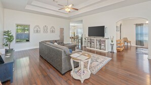 Un salon spacieux avec un canapé d’angle gris, un ventilateur de plafond, un téléviseur à écran plat et un meuble en bois.