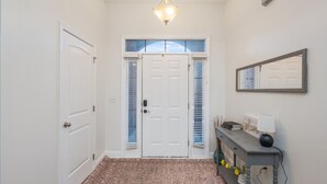 Un couloir avec une porte blanche, une fenêtre avec des stores, un luminaire et une console sur laquelle est posée une lampe.