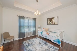 Um quarto com um sofá branco, uma poltrona, uma janela grande com cortinas, um lustre e um quadro na parede.