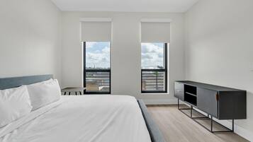 A modern bedroom with a large bed, a dark wooden cabinet, and a view of the city through two windows.