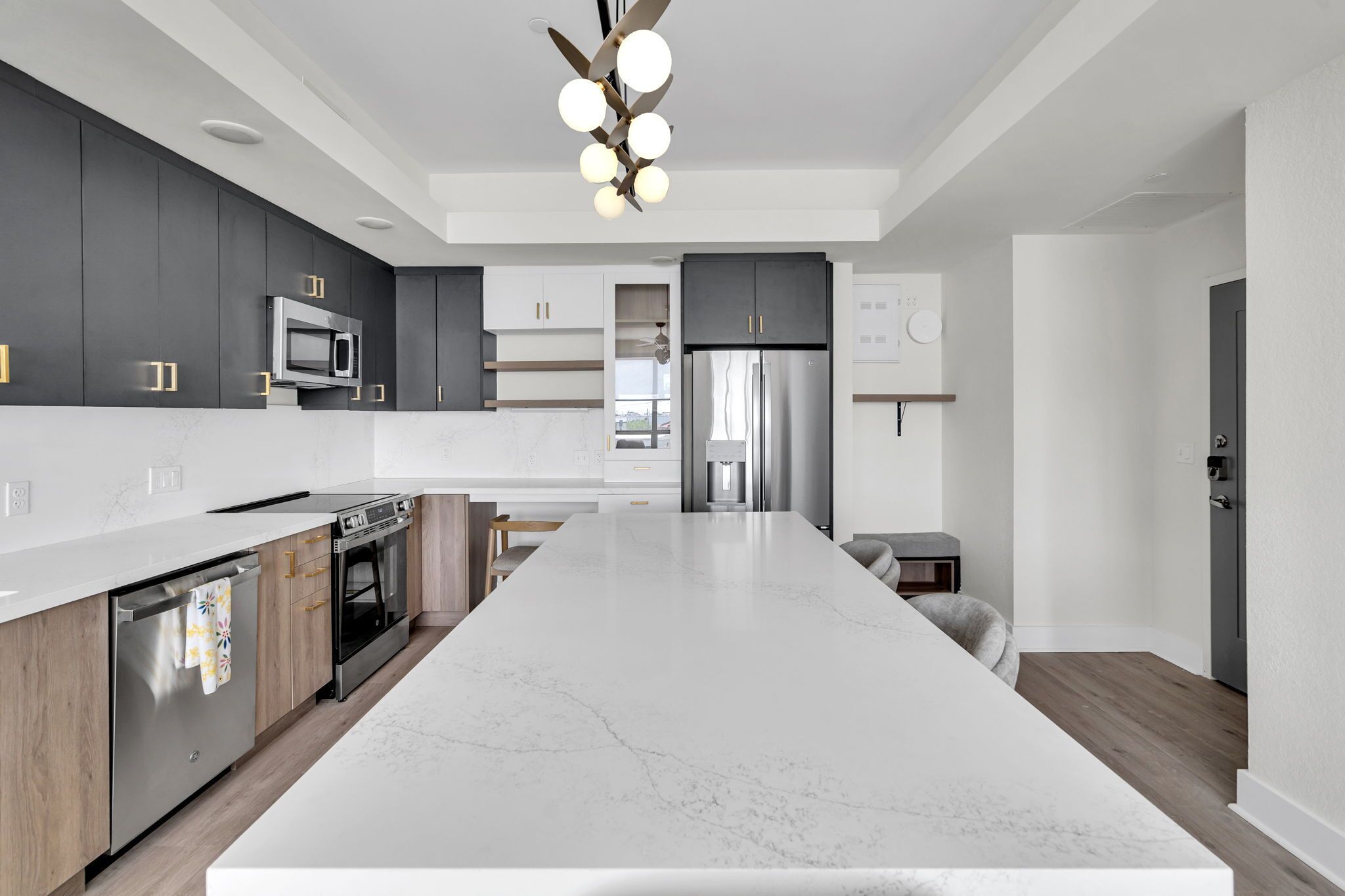 A modern kitchen with dark cabinets, a large marble island, stainless steel appliances, and a ceiling light fixture.