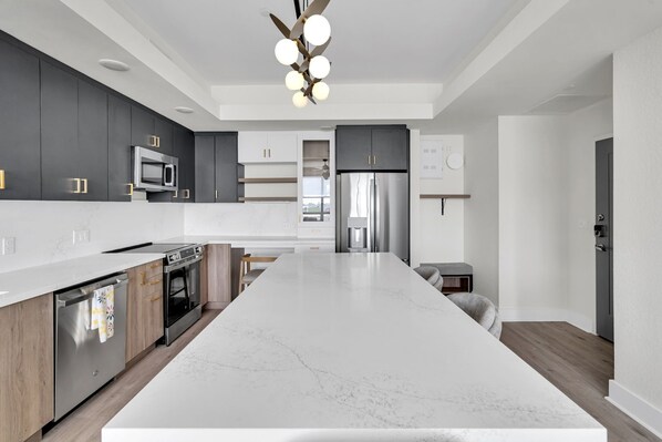 A modern kitchen with dark cabinets, a large marble island, stainless steel appliances, and a ceiling light fixture. - Latitude Apartments (Metairie)