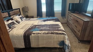 Une chambre à coucher avec un lit, une commode, une télévision et des fenêtres avec des rideaux.