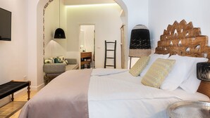 A hotel room with a large bed, a wooden headboard, a small table, a chair, a TV, and an arched doorway leading to another room. - Upscale 2-bedroom villa in the Venetian Harbour (Chania)