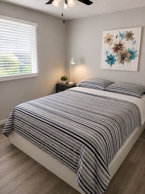A bedroom with a bed, a nightstand, a lamp, a ceiling fan, a window with blinds, and a floral painting on the wall.