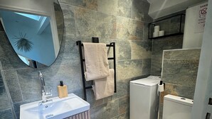 Une salle de bain moderne avec un mur en pierre, un miroir rond, un lavabo avec un meuble-vasque en bois, un porte-serviettes et des toilettes.