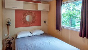Una cama individual con colchón y almohadas blancos, un espejo montado en la pared y una ventana con cortinas.
