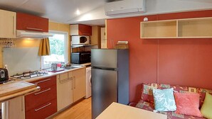 Cocina compacta con estufa a gas, microondas y refrigerador. Hay una ventana con cortinas, un lavamanos y un pequeño comedor con una mesa y sillas.