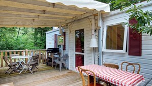 Una terraza techada al aire libre con mesa y sillas, una parrilla y una casa con contraventanas rojas.