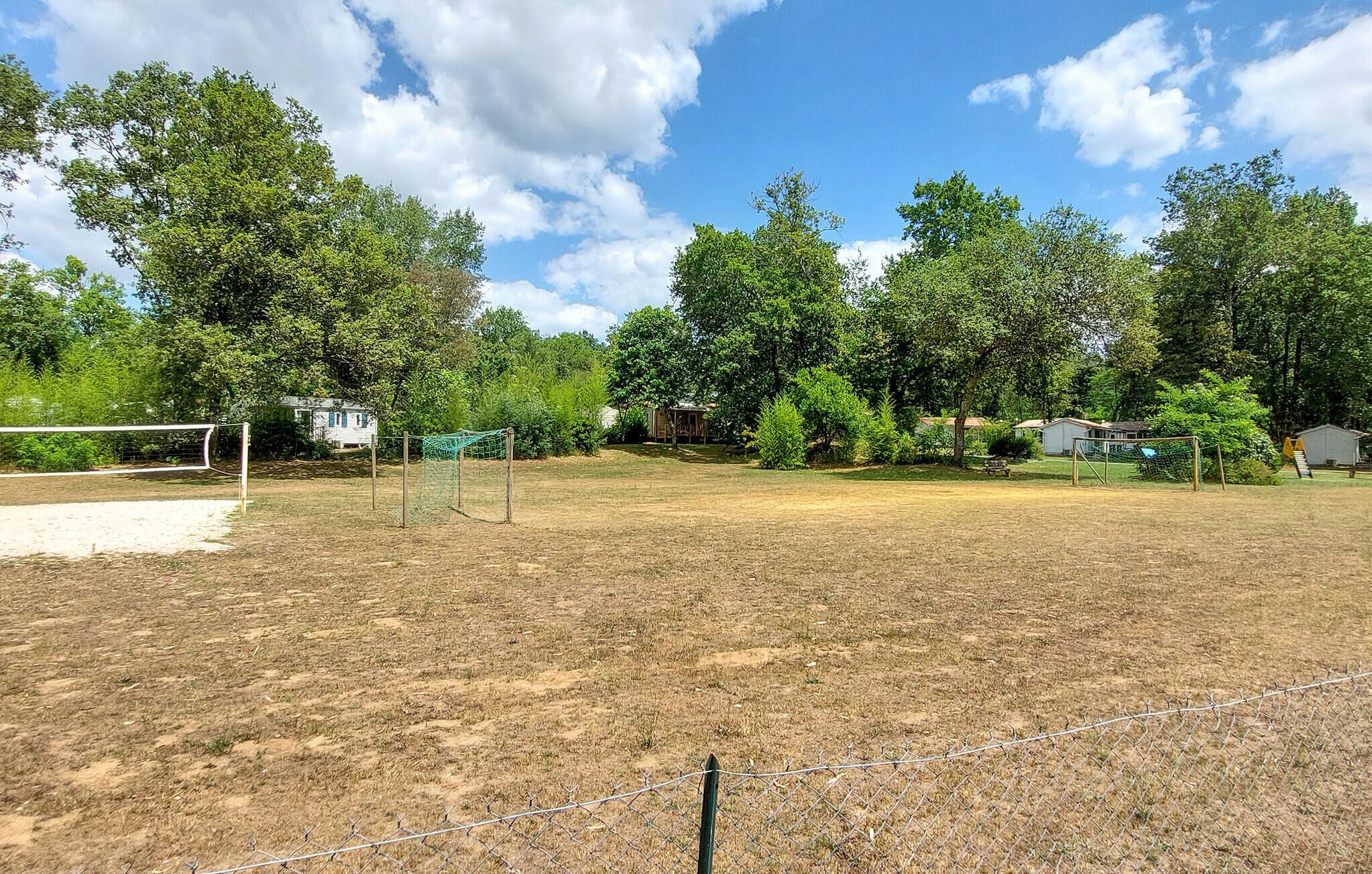 A fenced sports field with a volleyball net, surrounded by trees and houses in the background.