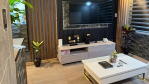 A modern living room with a flat-screen TV mounted on a dark wall, wooden slats, a white coffee table with remote controls, and decorative items. - Califnia Gardens Square (Mandaluyong)