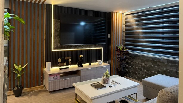 A modern living room with a flat-screen TV, wooden paneling, a stone accent wall, and a large window with blinds. - Califnia Gardens Square (Mandaluyong)