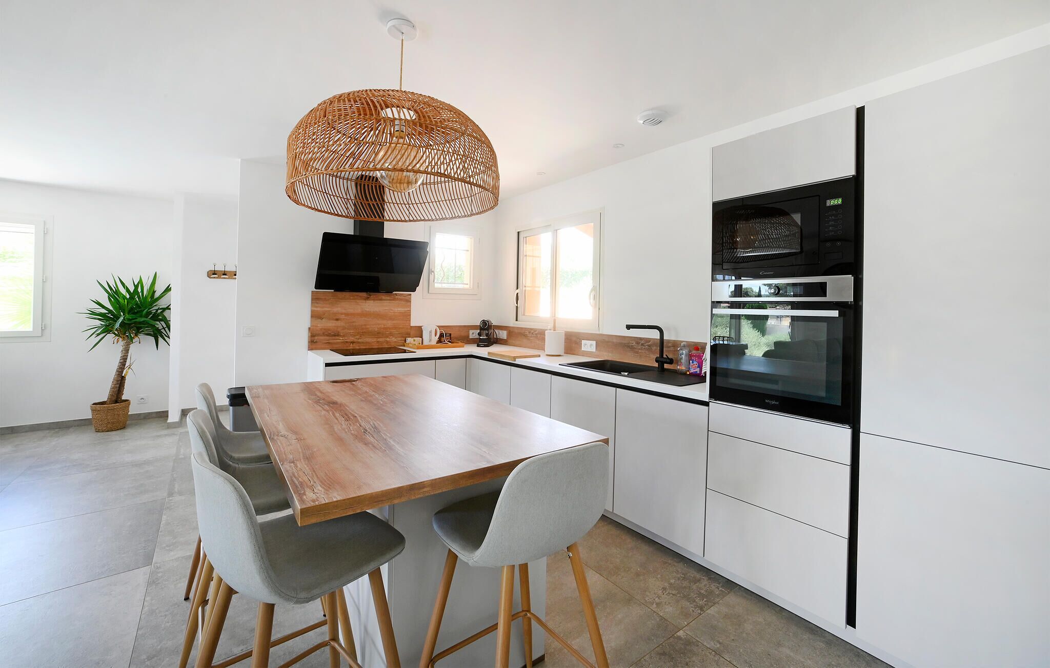 A modern kitchen with white cabinetry, a wooden dining table, grey chairs, a woven pendant light, and a built-in oven.