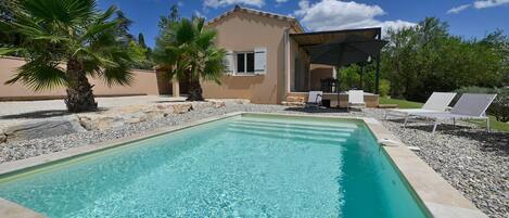A rectangular swimming pool with clear water, surrounded by a pebble border. There are lounge chairs and a covered outdoor seating area with a table and chairs. Palm trees are visible, and a building with a beige facade and white shutters is in the background.